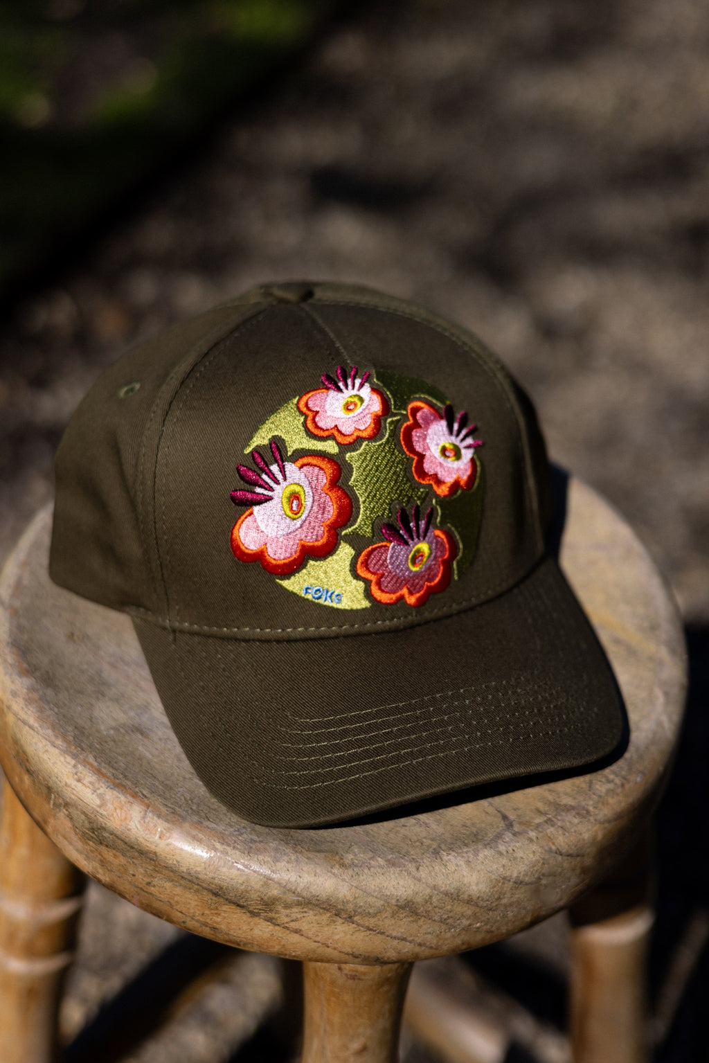 Olive FOKE cotton trucker cap with colourful floral embroidery, shown from the front on a wooden stool.