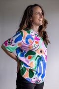 A woman wearing a colourful abstract-print FOKE tee, smiling and looking to the side against a grey studio background.