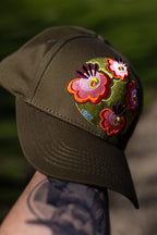 Olive FOKE trucker cap held outdoors, showing the curved peak and pink, orange and green floral embroidery.