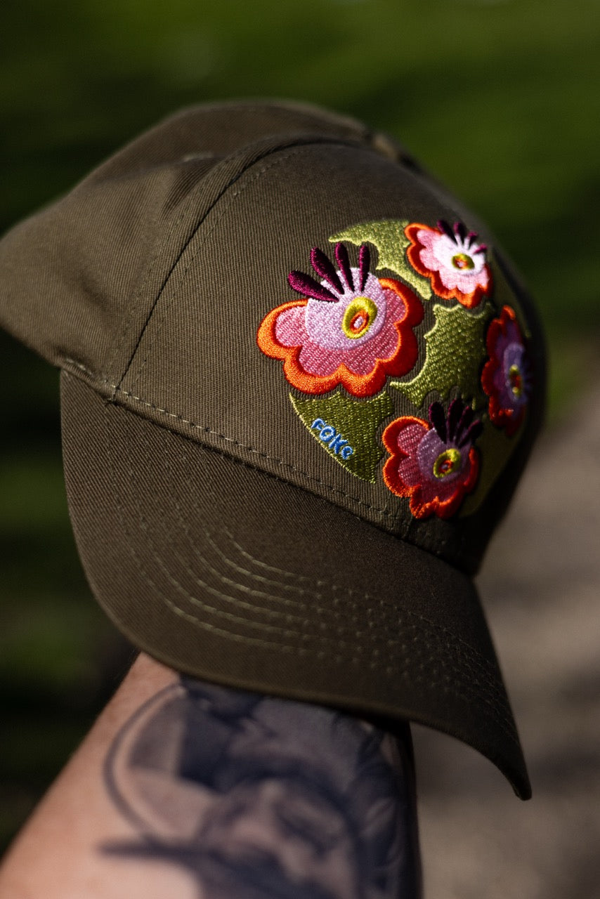 Olive FOKE trucker cap held outdoors, showing the curved peak and pink, orange and green floral embroidery.