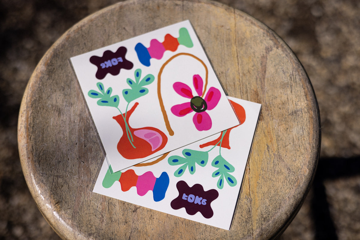 Two FOKE temporary tattoo sheets with colourful flowers, a vase and abstract shapes on a wooden stool.