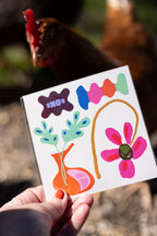 Hand holding one FOKE temporary tattoo sheet, showing the pink flower, orange vase and colourful graphic designs.