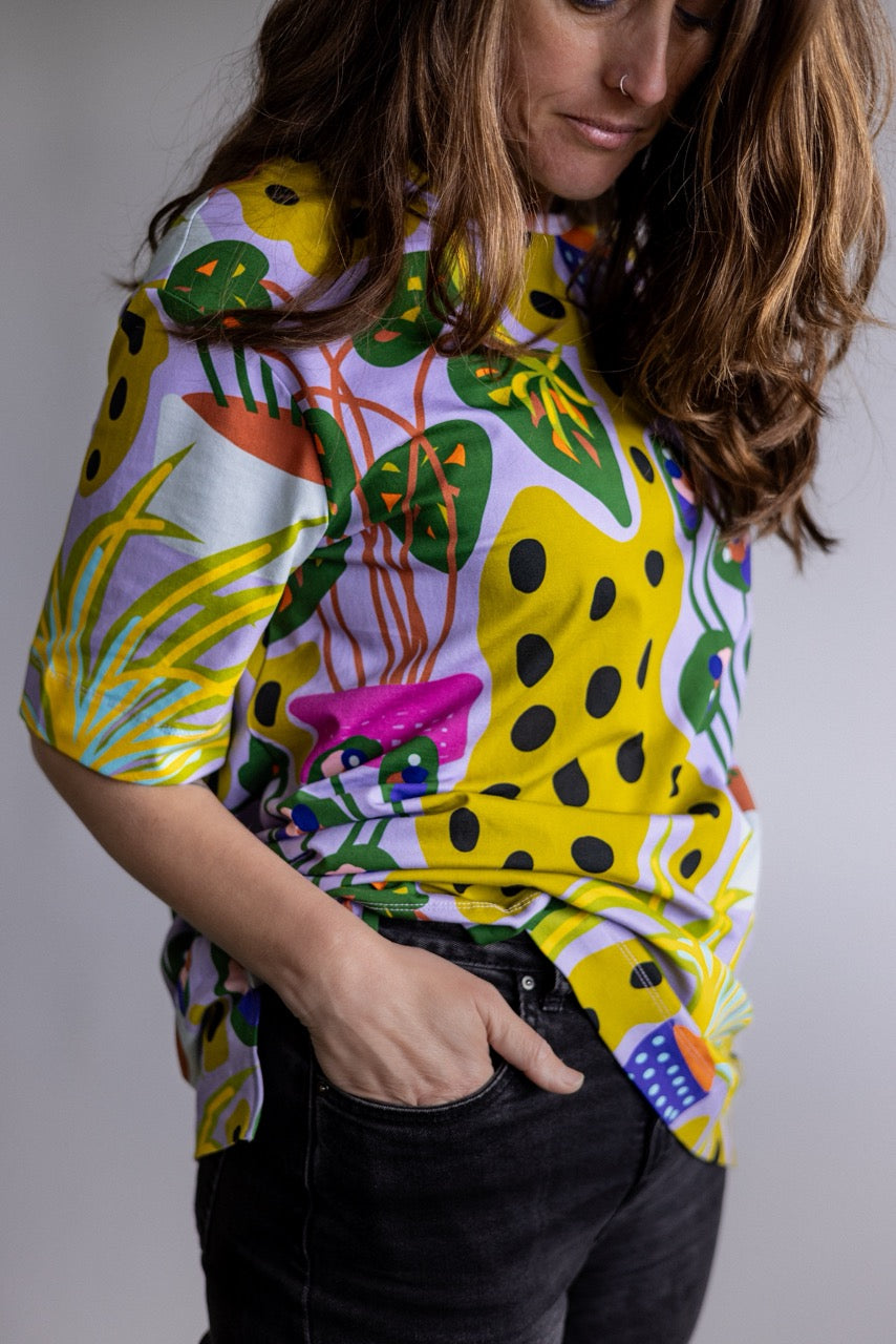 A woman wearing a short-sleeve high-neck top with a vibrant, multicolored abstract botanical print featuring yellow, green, pink, and black shapes on a light background, paired with black jeans.
