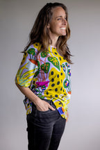 A woman wearing a vibrant yellow short-sleeve top with a colorful abstract botanical pattern, paired with black jeans, posing against a neutral gray studio background.