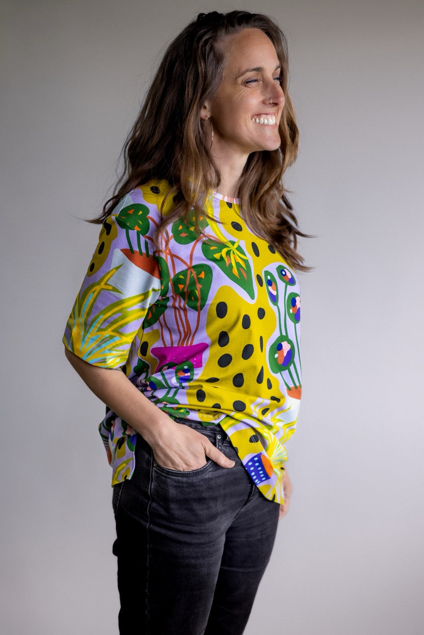 A woman wearing a vibrant yellow short-sleeve top with a colorful abstract botanical pattern, paired with black jeans, posing against a neutral gray studio background.