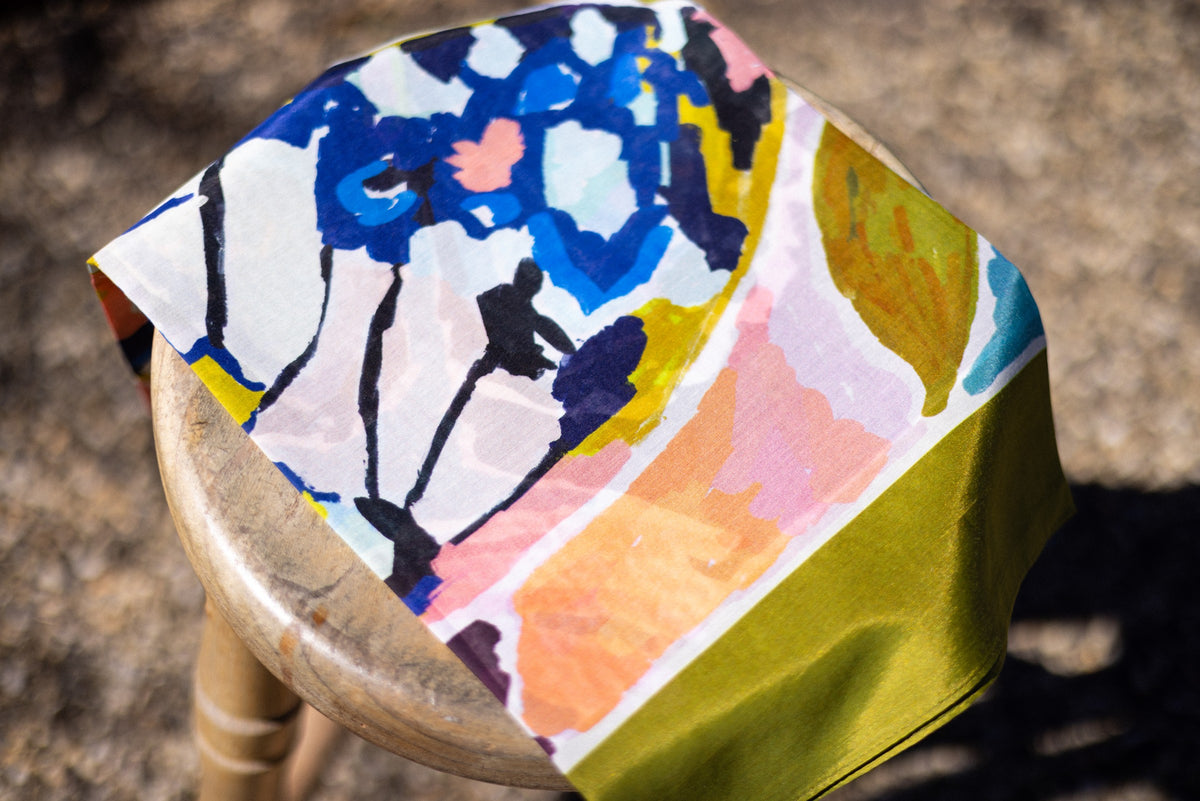 Folded Wild Reverie scarf on a wooden stool, showing blue butterfly wing details, pink brushwork and its olive border.