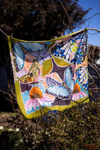 Wild Reverie cotton–silk scarf hanging outdoors, with blue butterflies, pink blooms and an olive border.