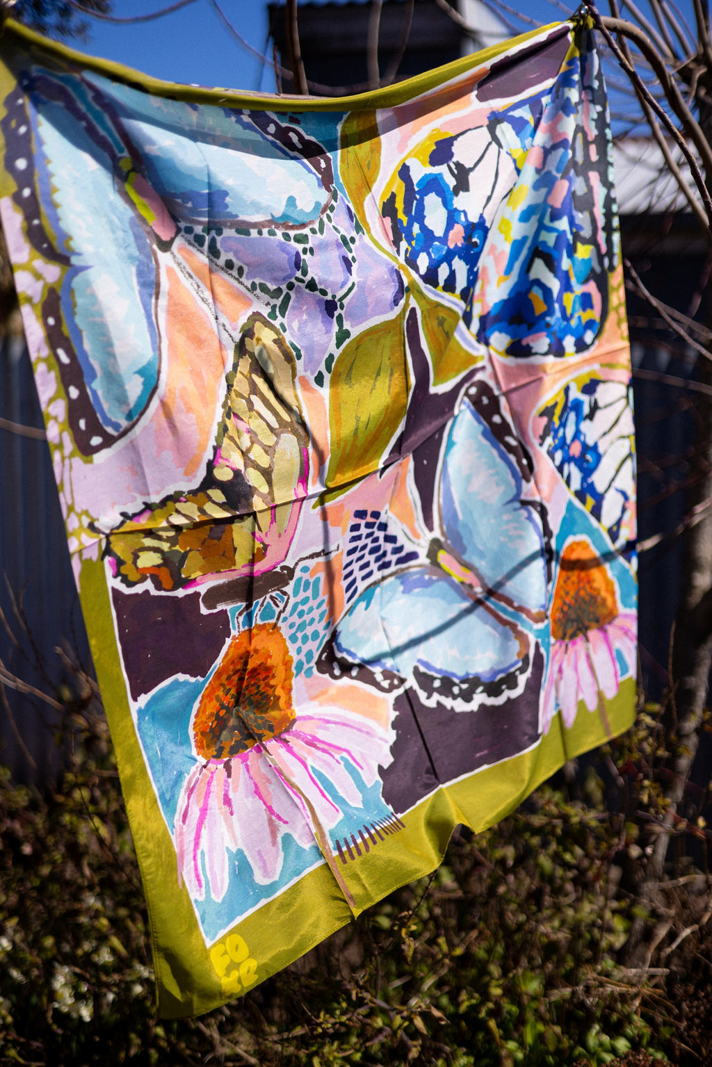 Wild Reverie scarf in sunlight, showing its sheer drape, large blue butterflies, pink flowers and olive border.
