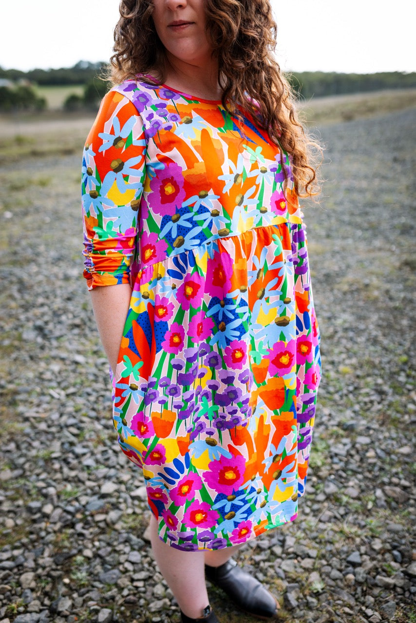 Person wearing a colorful abstract floral dress standing on a gravel path with a natural background. Colourful pattern detail — wearable art clothing by FOKE.