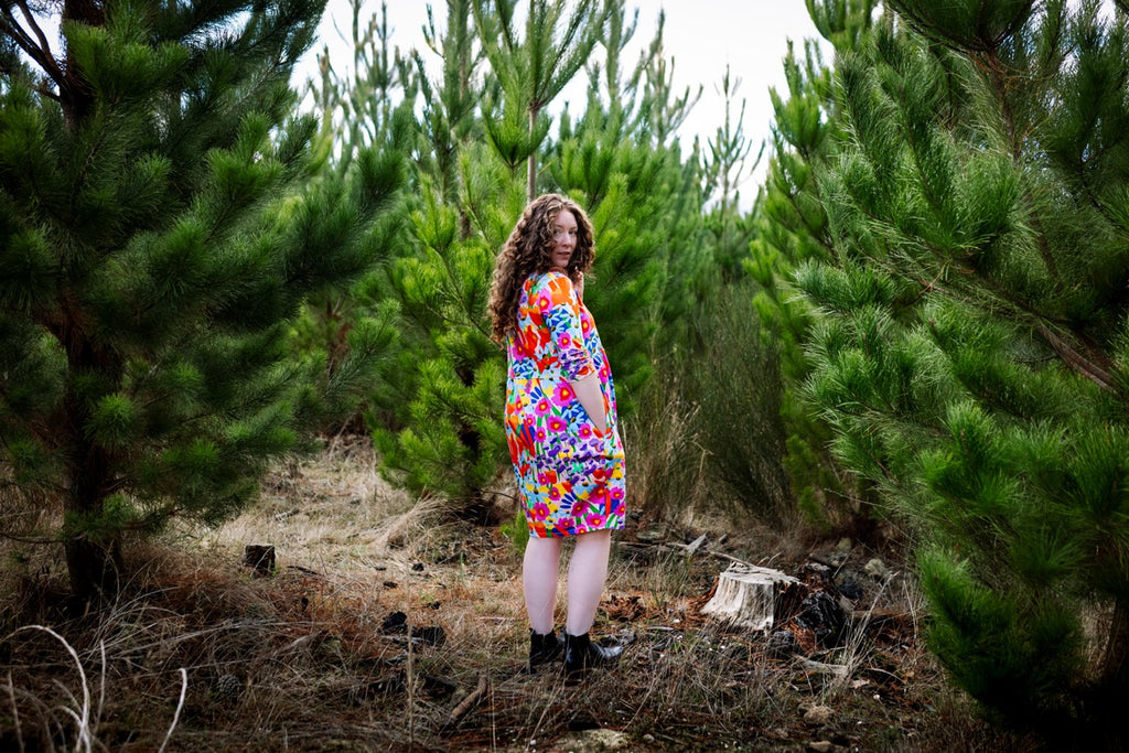 Person wearing a colorful dress standing among pine trees. Playful print detail — expressive Australian design fashion.