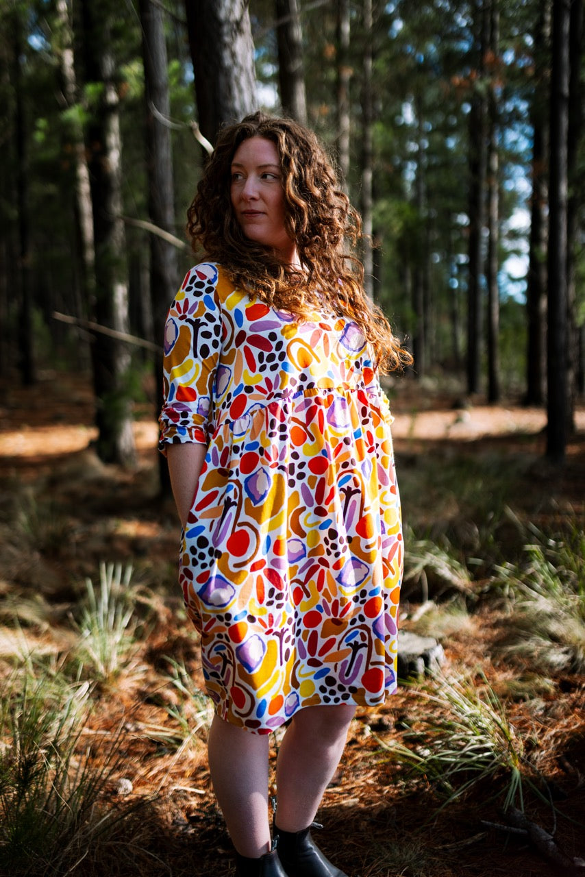 Woman wearing a colorful dress standing in a forest. Inclusive fit hand-drawn print dress for bold women shaping bold stories.