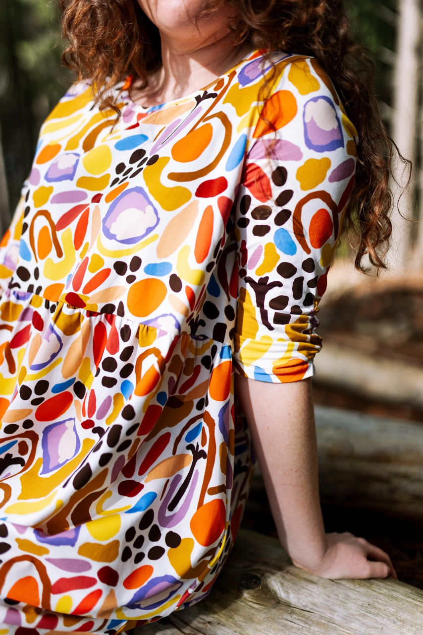 Person wearing a colorful abstract patterned dress sitting outdoors. High-quality wearable art clothing designed for everyday comfort.